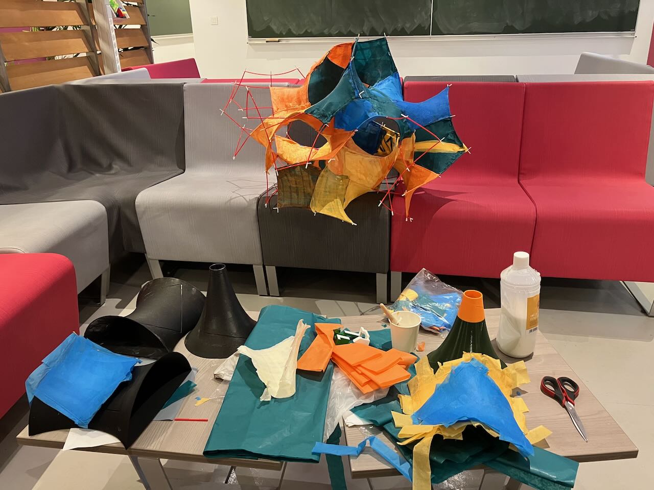the common area during construction. There is a table covered in paper, scissors, 3D printed models, and glue. In the back, there is the hyperbolic piniata  