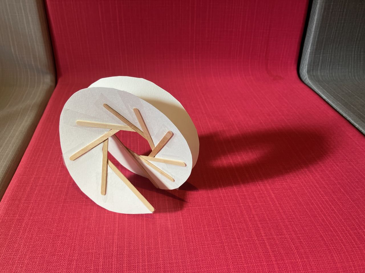 A small piece of a helix, made in paper supported by popsicle sticks. It is bathed in sunlight, casting a circular shadow